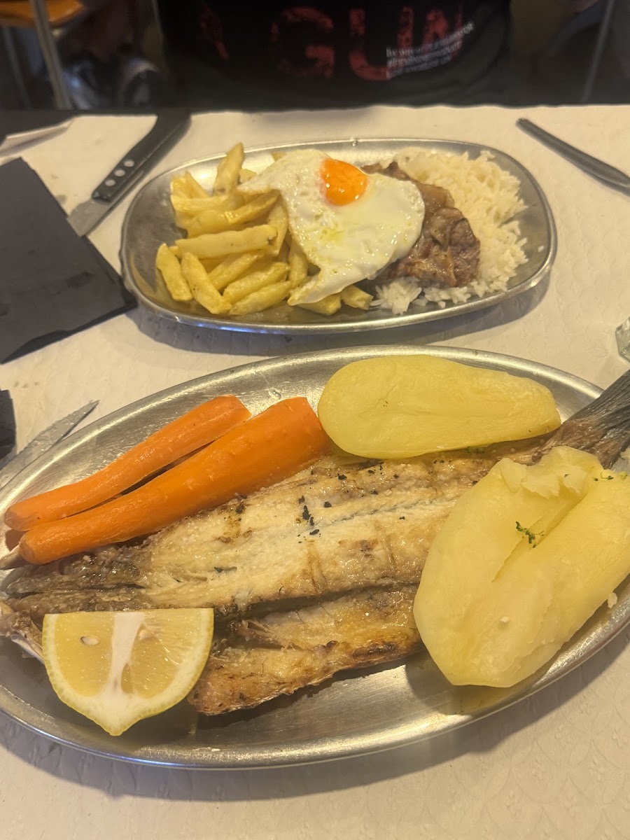 Travessa oval de metal com peixe frito, batata cozida e cenoura, ao lado de outra com arroz, carne e ovo estrelado