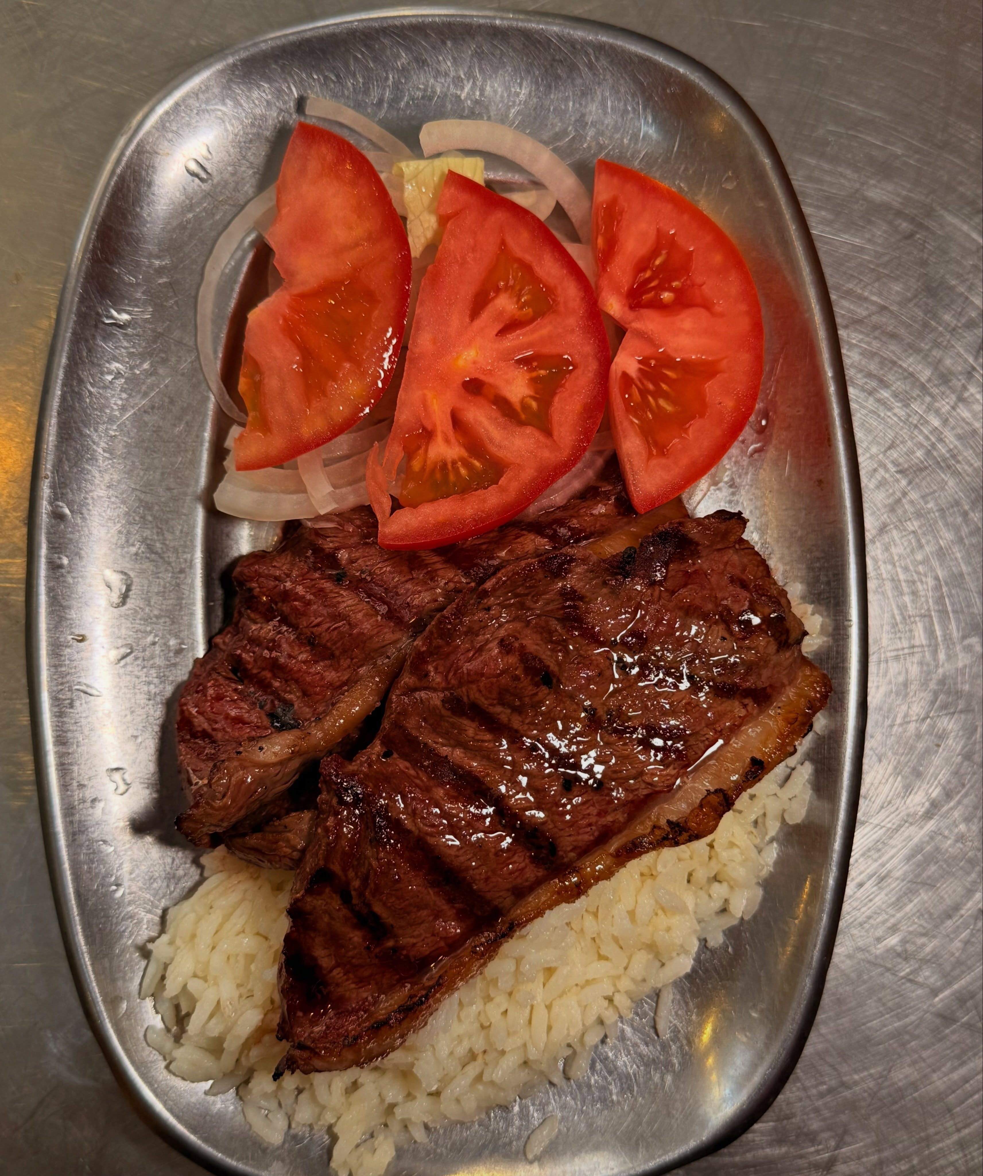 Bife grelhado com arroz branco, tomate e cebola numa travessa de metal