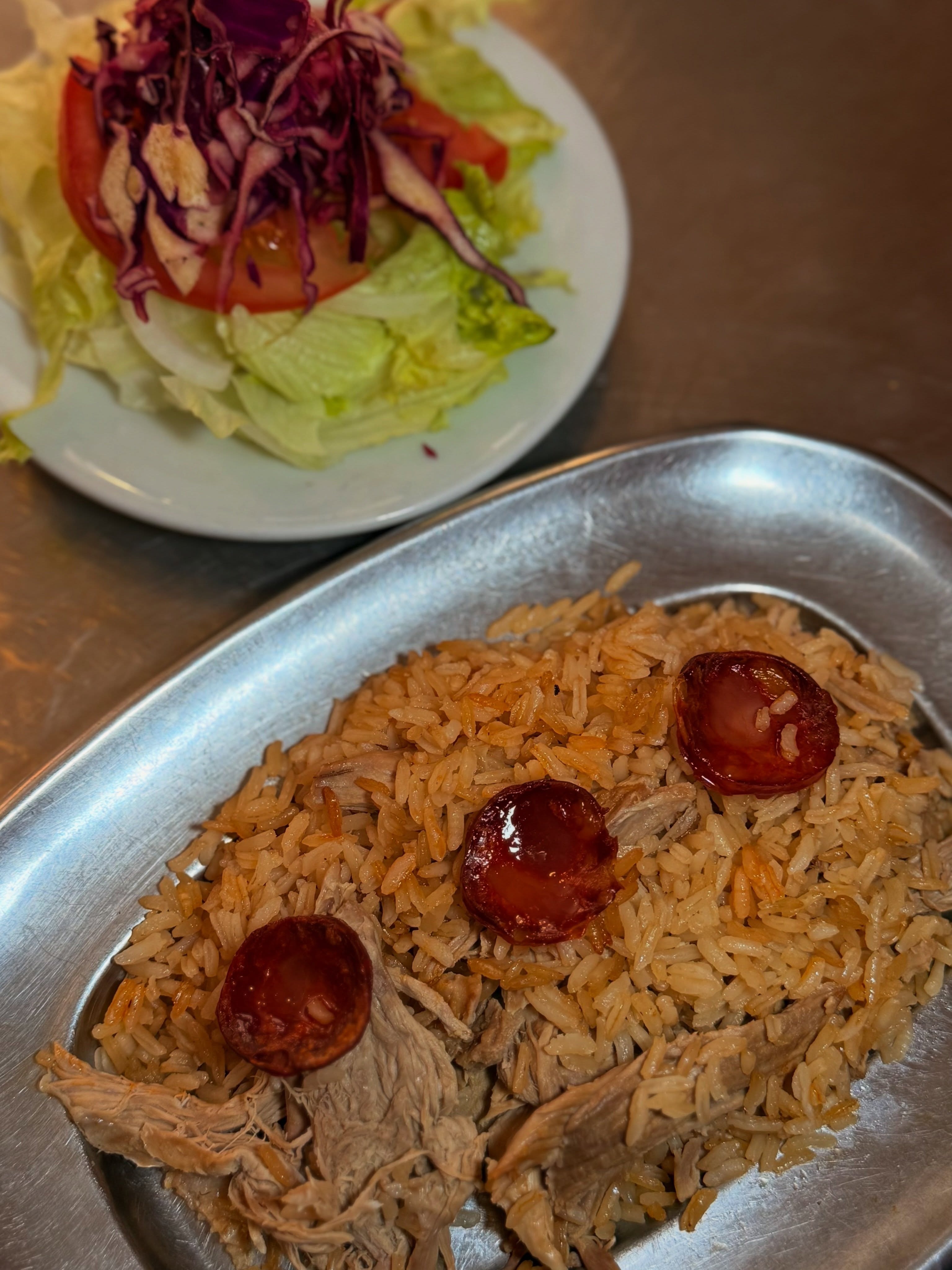 Arroz com carne desfiada e chouriço numa travessa de alumínio, com salada ao fundo